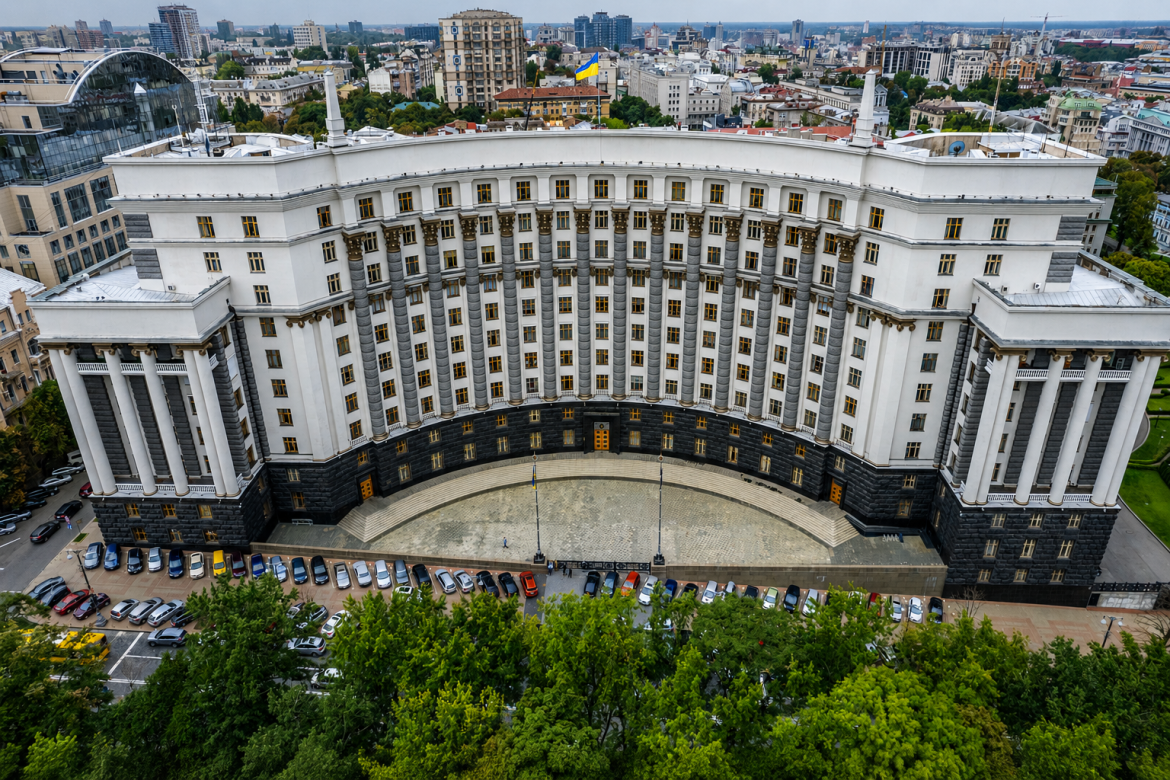 Аерофотознімок масивної урядової будівлі напівкруглої форми з колонами та українським прапором у центрі Києва в оточенні зелених дерев і паркування